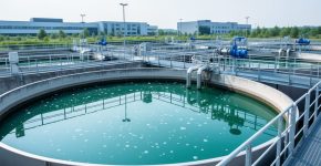 Modern wastewater treatment facility with aeration tanks, settling basin water, and industrial pipes under a bright sky
