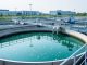 Modern wastewater treatment facility with aeration tanks, settling basin water, and industrial pipes under a bright sky