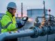 Technician using an infrared methane detector near pipelines and valve equipment at an industrial facility during dawn conditions.